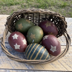 May include: A wicker basket filled with six hand-painted Easter eggs. The eggs are decorated with stars, stripes, and a variety of colours, including red, green, and gold.