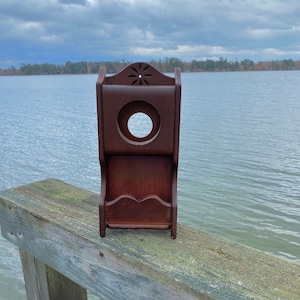 May include: A dark brown wooden wall mounted shelf with a decorative cutout in the front. The shelf has a small shelf on the bottom and a larger shelf on the top. The shelf is mounted on a wooden railing.