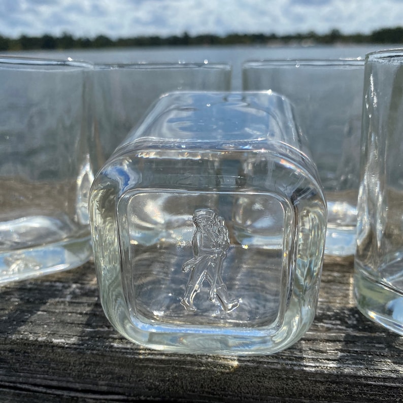May include: Four clear glass tumblers with a square base and a raised design of a person walking on the front of the glass.