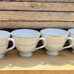 May include: Four white teacups with a silver trim and handle. Each cup has a silver band around the middle and sits on a small, white pedestal base. The cups are arranged in a row.