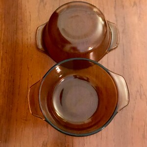 Puede incluir: Dos cuencos de vidrio color ámbar con asas. Los cuencos son redondos con un borde ligeramente ensanchado y están hechos de vidrio transparente y resistente al calor. Un cuenco está boca abajo, revelando su base. Los cuencos están sobre una superficie de madera.