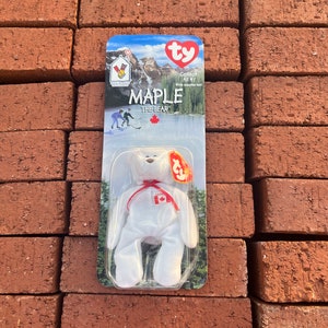 Könnte beinhalten: Ein weißes Plüschtier namens "Maple" aus der Ty Beanie Baby-Kollektion. Der Bär hat ein rotes Band und eine kanadische Flagge. Die Verpackung zeigt einen See und Berge. Der Text lautet "Canada" und "Maple the Bear".