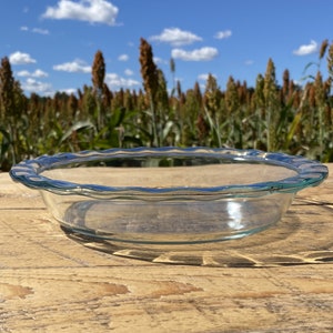 Puede incluir: Un molde para tarta de vidrio transparente con un borde ondulado y festoneado. El plato está sobre una superficie de madera desgastada, con un campo de plantas altas y un cielo azul con nubes en el fondo. El molde para tarta está vacío.