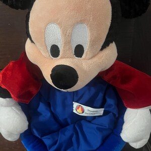 May include: A plush Mickey Mouse character with a black head and ears, a tan face, and a black nose. It wears a blue outfit with a red cape. The Disneyland logo is visible on the outfit.