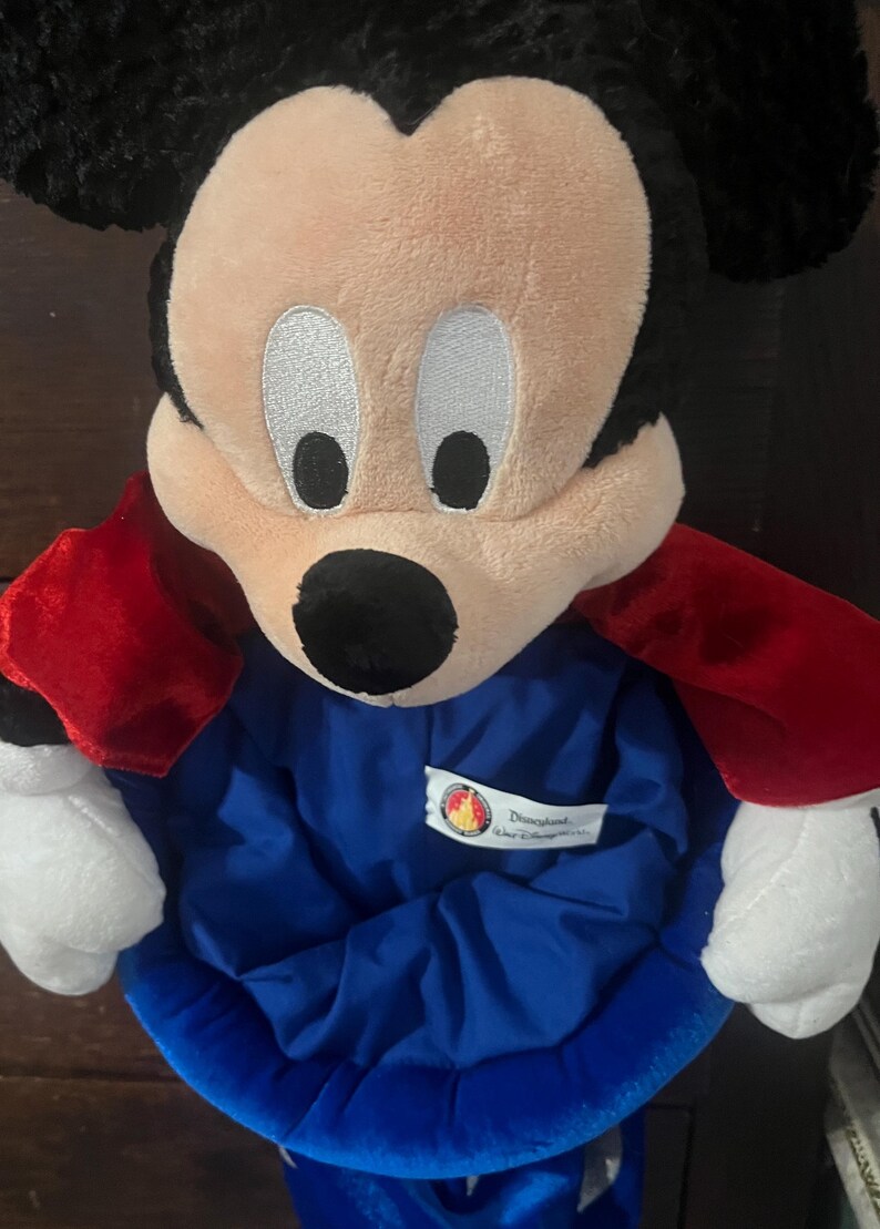 May include: A plush Mickey Mouse character with a black head and ears, a tan face, and a black nose. It wears a blue outfit with a red cape. The Disneyland logo is visible on the outfit.