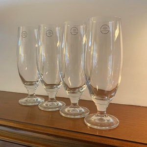 May include: Four clear, stemmed beer glasses with a rounded bowl and a flared rim. Each glass has a circular logo near the top. The glasses are displayed on a wooden surface.