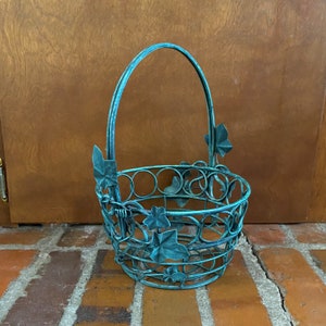 Puede incluir: Una cesta de alambre de metal azul verdoso con asa y detalles de hojas de hiedra. La cesta está diseñada para guardar objetos pequeños.