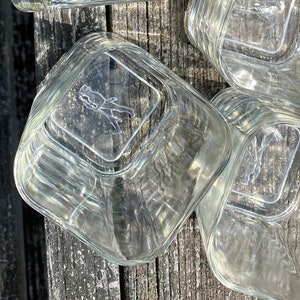 May include: A set of four clear glass square containers with a raised design on the bottom. The design features a stylized figure of a person.