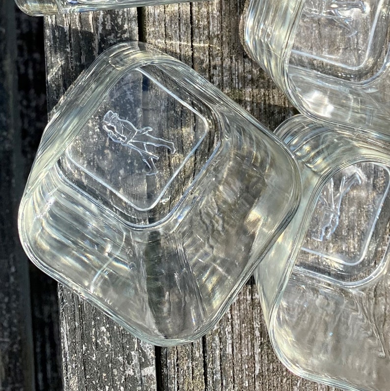 May include: A set of four clear glass square containers with a raised design on the bottom. The design features a stylized figure of a person.