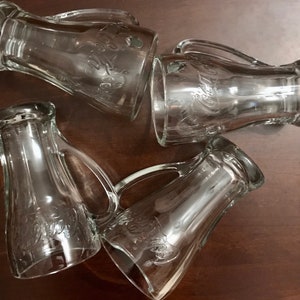 Op de afbeelding: Vier heldere glazen bekers met handvatten. Elke beker heeft het woord "Coca-Cola" in het glas gegraveerd.