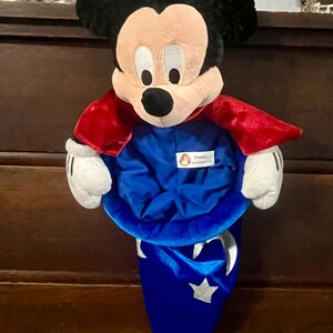 May include: A Mickey Mouse plush toy wearing a blue wizard hat and a red cape. The plush has black ears, a tan face, and white gloves. The hat has a silver crescent moon and star design.