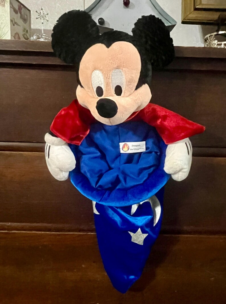 May include: A Mickey Mouse plush toy wearing a blue wizard hat and a red cape. The plush has black ears, a tan face, and white gloves. The hat has a silver crescent moon and star design.