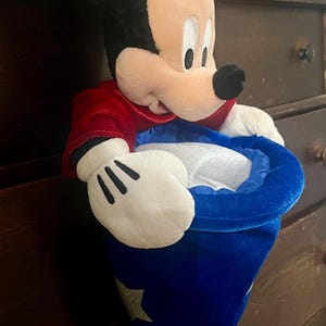 May include: A plush Mickey Mouse toy wearing a red jacket and a blue wizard's hat with silver stars. The toy has black ears, a white face, and white gloves. The hat is open, revealing a white interior.
