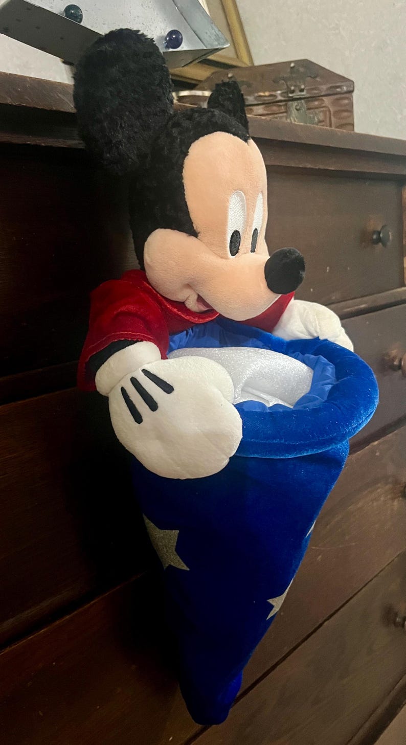 May include: A plush Mickey Mouse toy wearing a red jacket and a blue wizard's hat with silver stars. The toy has black ears, a white face, and white gloves. The hat is open, revealing a white interior.