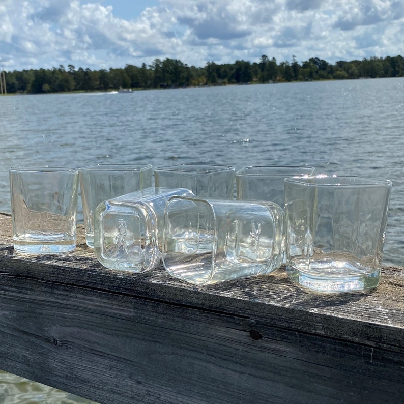 May include: A set of eight clear glass tumblers with a square shape. The glasses are arranged in a row on a wooden surface.
