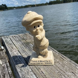 May include: A beige figurine of a cartoon character wearing a cap, sweater, and scarf. The figurine is standing on a square base with the text "There's A Word For A Person Like You...TERRIFIC". The background is a lake.