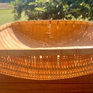 May include: A square, woven basket made of light brown bamboo. The basket has a rounded rim and a shallow depth. The weave is tight and even, creating a sturdy container. The basket is sitting on a wooden surface.