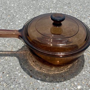 Puede incluir: Una cacerola de vidrio marrón con tapa y asa a juego. La batería de cocina está hecha de vidrio transparente, lo que permite ver el contenido. La sartén tiene aproximadamente 25 cm de diámetro.
