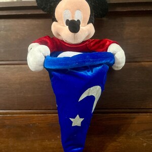 May include: A Mickey Mouse plush toy with a red shirt and black ears. The plush is holding a blue velvet hat with a silver crescent moon and star design. The hat is open, revealing a white interior.