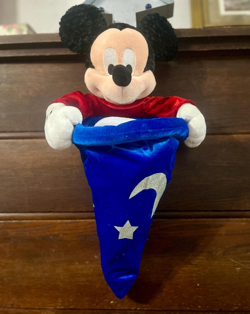 May include: A Mickey Mouse plush toy with a red shirt and black ears. The plush is holding a blue velvet hat with a silver crescent moon and star design. The hat is open, revealing a white interior.