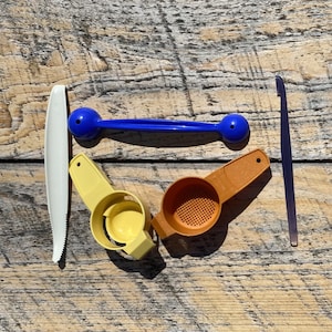 以下が含まれることがあります： 白いギザギザのナイフ、青い泡立て器、黄色の卵セパレーター、茶色のストレーナー、紫色のスパチュラなど、さまざまなキッチン用品。木製の表面に配置されています。