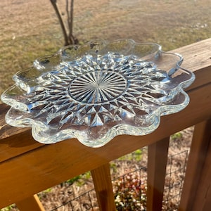 May include: Clear glass serving plate with a scalloped edge and a detailed, starburst-like pattern in the center. The plate is resting on a wooden surface, catching the sunlight.