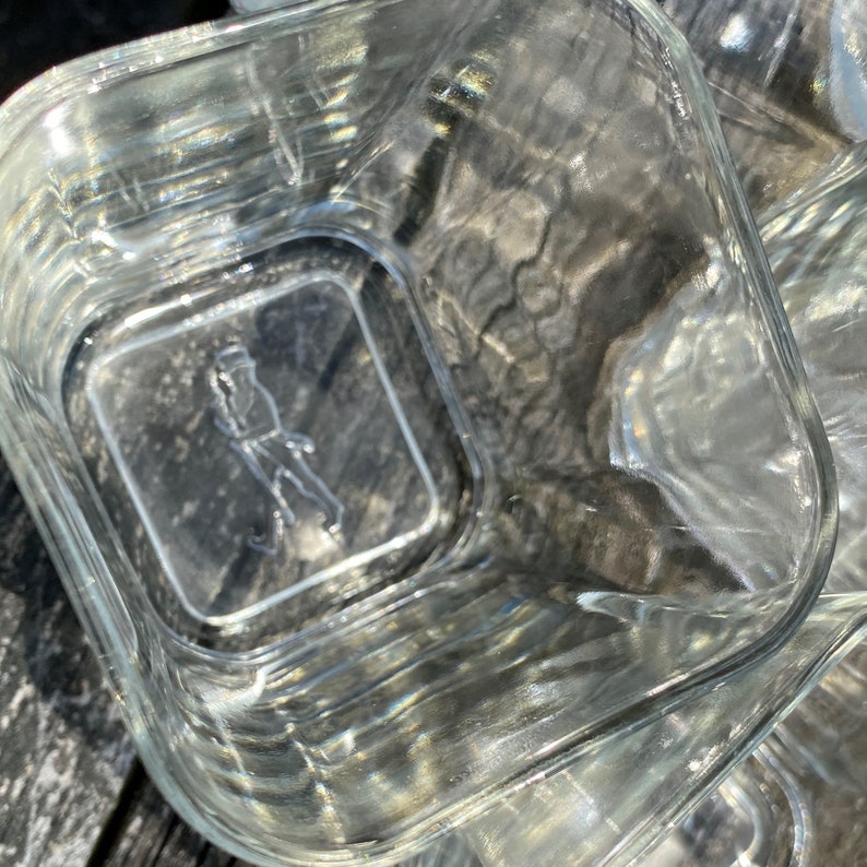 May include: A clear glass square container with a raised design of a person walking on the bottom. The container is empty and appears to be new.