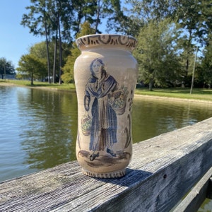 Puede incluir: Un jarrón de cerámica con un diseño pintado a mano de una mujer que lleva una cesta de fruta. El jarrón es beige con detalles azules y verdes.