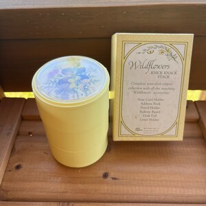 May include: A yellow cylindrical stackable container with a clear lid featuring a floral print, accompanied by its original rectangular box. The box is labeled Wildflowers Knick Knack Stack and lists matching desk accessories including a note card holder.