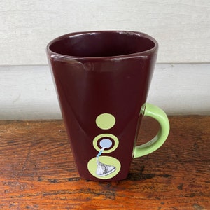 May include: A maroon ceramic mug with a green handle. The mug has a design of a chocolate kiss with a green, blue, and white circle pattern.