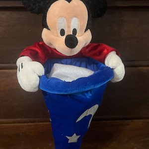 May include: A plush Mickey Mouse figure wearing a red shirt and holding a blue wizard's hat with white stars and crescent moons. The hat is open, revealing a white object inside. The figure has black ears, a tan face, and white gloves.