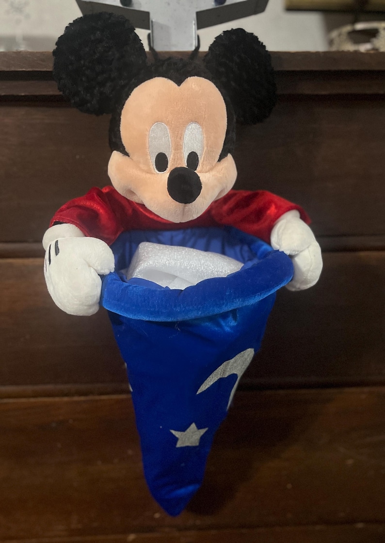May include: A plush Mickey Mouse figure wearing a red shirt and holding a blue wizard's hat with white stars and crescent moons. The hat is open, revealing a white object inside. The figure has black ears, a tan face, and white gloves.