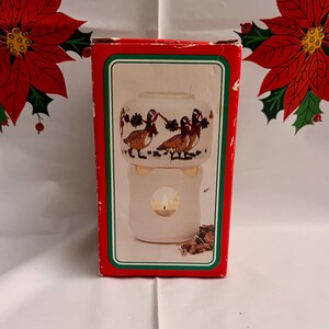 May include: A decorative candle holder in a red box with a green border. The candle holder is white with a duck design. The box has a poinsettia design on the top and bottom.