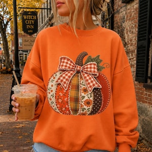 Op de afbeelding: Een oranje sweater met ronde hals en een grote, centrale pompoenafbeelding. De pompoen is samengesteld uit diverse stoffen patches met bloemen-, ruit- en stippenpatronen, bekroond met een grote rood-witte gingham stoffen strik.