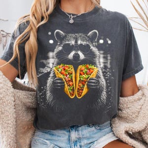 May include: Dark gray t-shirt featuring a raccoon wearing sunglasses and holding two tacos. The raccoon is in a detailed graphic design. The tacos are filled with meat, tomatoes, lettuce, and cheese. The shirt is a casual wear item.