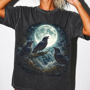 May include: A charcoal gray short-sleeve t-shirt featuring a graphic print of three black ravens perched on tree branches against a backdrop of a large, bright full moon, a starry night sky, and swirling clouds in shades of blue and white.