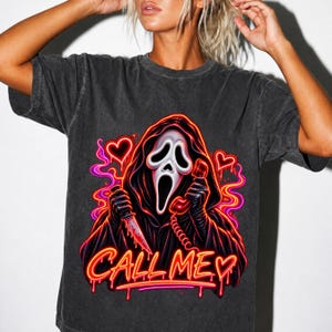 Puede incluir: Una camiseta de manga corta gris carbón con un gráfico de una figura encapuchada con máscara blanca que sostiene un cuchillo y un teléfono rojo. El diseño incluye contornos de neón rosa y naranja, corazones y el texto CALL ME en fuente goteante.