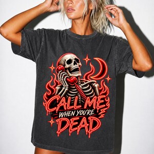Puede incluir: Una camiseta de manga corta en color gris carbón con un gráfico de un esqueleto sosteniendo un auricular de teléfono rojo. El diseño incluye llamas rojas estilo neón, una luna creciente y el texto "CALL ME WHEN YOU'RE DEAD" en rojo.