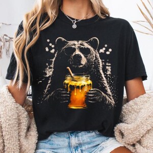 May include: Black t-shirt featuring a bear wearing sunglasses and holding a jar of honey with a straw. The bear graphic is detailed and the honey jar is a vibrant yellow. The shirt is a casual style.