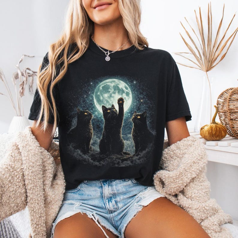 May include: Black t-shirt featuring a graphic of three black cats in front of a full moon. One cat is reaching up towards the moon. The cats are sitting on a cloud-like formation. The background is a starry night sky.