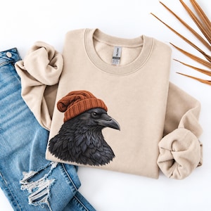 May include: A beige crewneck sweatshirt featuring a graphic of a black raven wearing a textured orange knit beanie. The sweatshirt has long sleeves and a ribbed collar, with a Gildan brand tag visible at the inner neckline, paired with blue jeans.