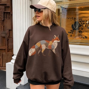 May include: A dark brown crewneck sweatshirt featuring a large graphic of a fox on the front. The fox is composed of various patchwork-style patterns, including floral prints, checkered designs, and solid colors in shades of orange, brown, and cream.