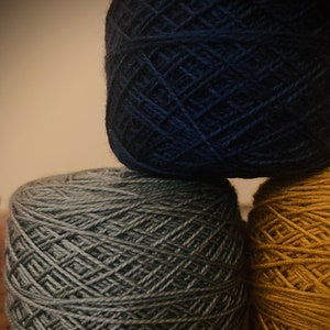 May include: Three balls of yarn stacked on top of each other. The top ball is dark blue, the middle ball is a muted green, and the bottom ball is a bright yellow.