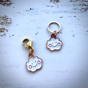 May include: Two gold-tone cloud charms with pink cheeks and a smiling face. One charm has a lobster clasp and the other has a jump ring.