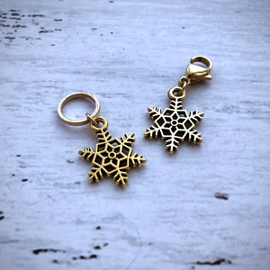 May include: Two gold snowflake charms with a ring and a lobster clasp. The charms are on a white wooden surface.