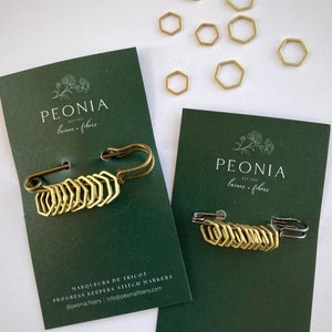 May include: Two green cards with the text "PEONIA laines - fibers EST 2021" and a floral design. Each card features a gold safety pin with a chain of hexagonal stitch markers. The text "MARQUEURS DE TRICOT PROGRESS KEEPERS STITCH MARKERS @peonia.fibers | info@peoniafibers.com" is on the bottom of the card on the left.