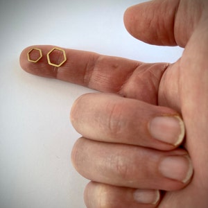 May include: Two gold hexagon shaped earrings, one slightly larger than the other, are shown on a finger.