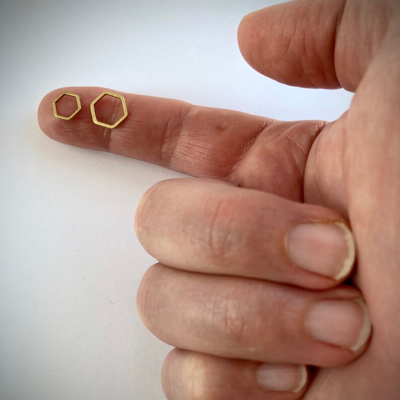 May include: Two gold hexagon shaped earrings, one slightly larger than the other, are shown on a finger.