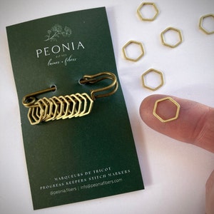 May include: A set of gold-toned hexagonal stitch markers, with a safety pin attached to a chain of markers. The markers are labeled "Marqueurs de Tricot" and "Progress Keepers Stitch Markers" with the brand name "Peonia laines fibers" and website information.
