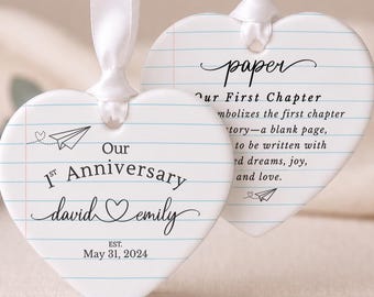 May include: Two heart-shaped ceramic ornaments with a white lined paper pattern and white satin ribbons. The front features the text Our 1st Anniversary, the names david and emily with a heart, and the date May 31, 2024. The back features text about paper.
